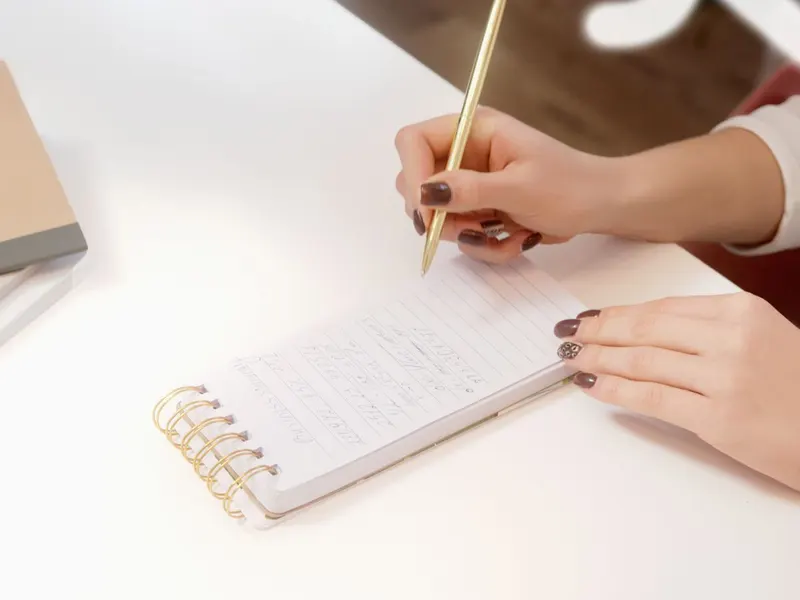 Mão anotando em um caderno de espiral sobre uma mesa branca